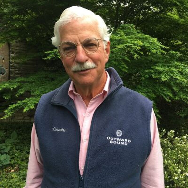 The image shows an older man with white hair, glasses, and a mustache. He is wearing a pink shirt and a blue vest with the 'Outward Bound' logo. He is standing in front of some green bushes. The man appears to be looking at the camera.